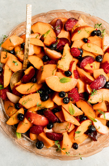 An overhead shot shows a summer fruit salad on a pink, etched glass platter with a brass serving spoon sticking out. The salad is topped with fresh sliced mint.