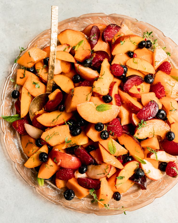 An overhead shot shows a summer fruit salad on a pink, etched glass platter with a brass serving spoon sticking out. The salad is topped with fresh sliced mint.