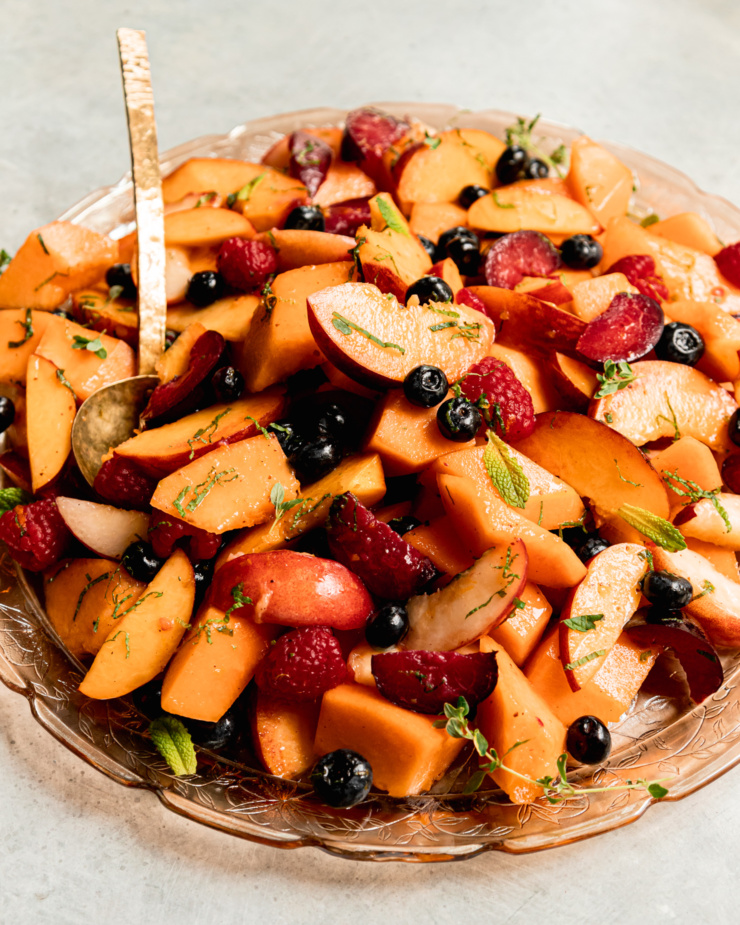 A 3/4 angle shot shows a summer fruit salad on a pink, etched glass platter with a brass serving spoon sticking out. The salad is topped with fresh sliced mint.
