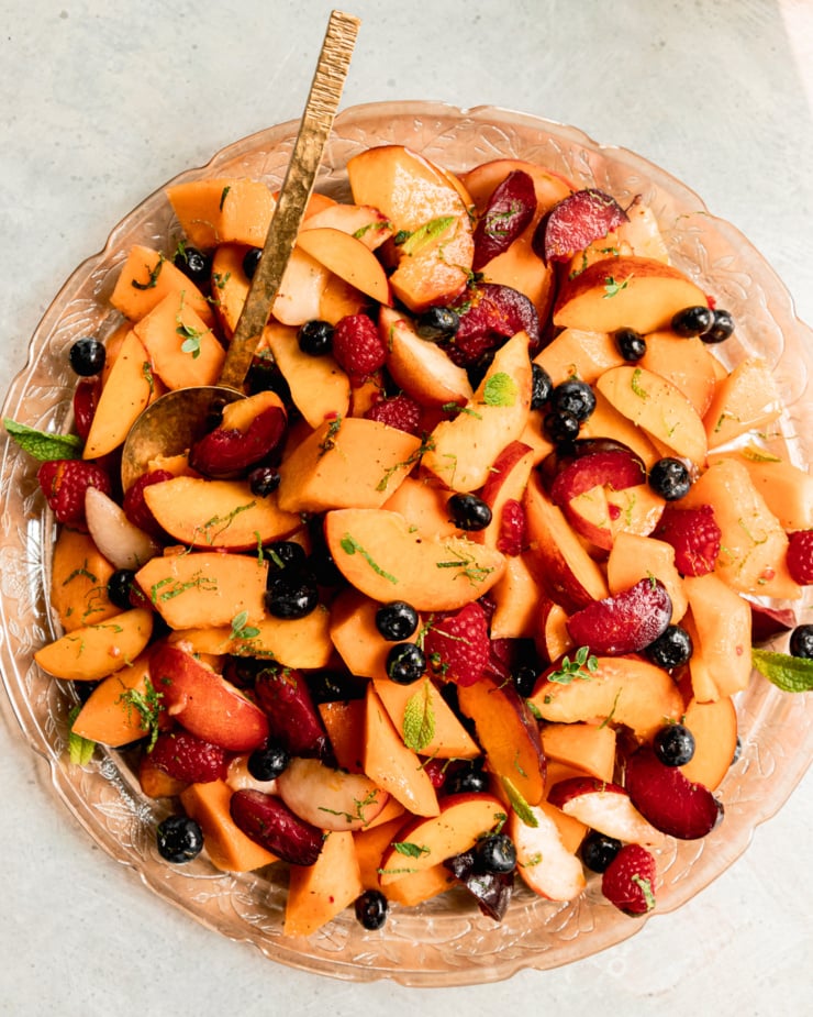 An overhead shot shows a summer fruit salad on a pink, etched glass platter with a brass serving spoon sticking out. The salad is topped with fresh sliced mint.