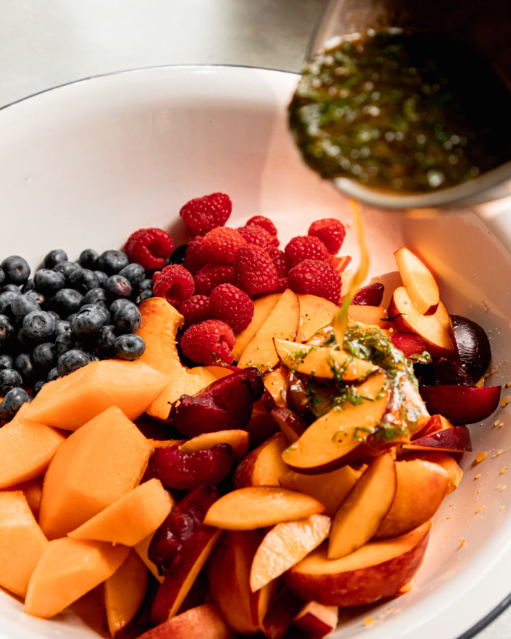 A 3/4 angle shot shows an orange mint dressing being poured over some fruit in a wide white bowl.