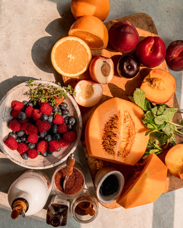 An overhead shot shows some fruit and fresh herbs on a worn wood cutting board and in a grey ceramic board. Olive oil, spices, maple syrup, and vanilla are seen nearby.