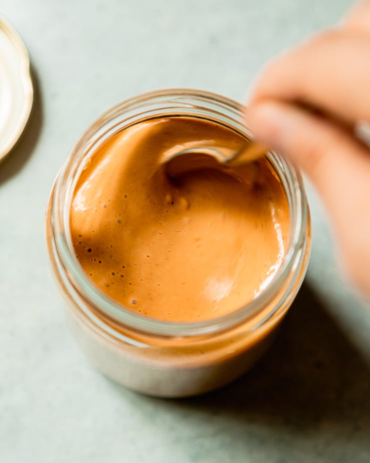 An overhead shot shows a hand using a spoon to stir up a jar of peanut sauce.