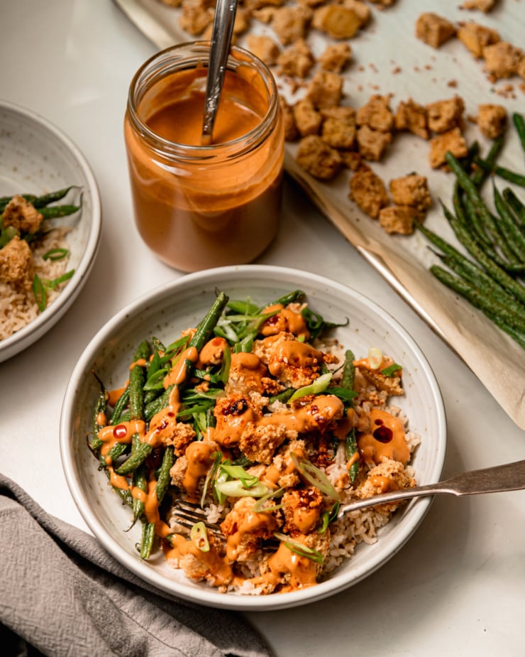 A slight 3/4 angle shot of a bowl of crispy peanut tofu and green beans over brown rice. The bowl is garnished with chili crisp and green onions. A jar of peanut sauce is nearby.