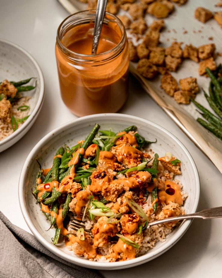 A slight 3/4 angle shot of a bowl of crispy peanut tofu and green beans over brown rice. The bowl is garnished with chili crisp and green onions. A jar of peanut sauce is nearby.