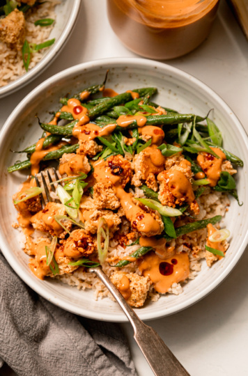 An overhead shot of a bowl containing cooked brown rice, crispy peanut tofu, and green beans. The bowl is topped with drops of chili crisp and sliced green onions. There are also extra drizzles of peanut sauce.