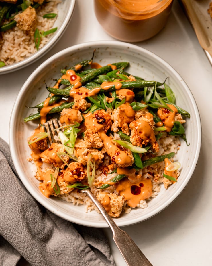 An overhead shot of a bowl containing cooked brown rice, crispy peanut tofu, and green beans. The bowl is topped with drops of chili crisp and sliced green onions. There are also extra drizzles of peanut sauce.