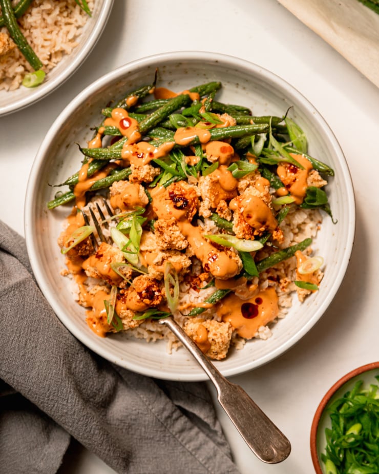 An overhead shot of a bowl containing cooked brown rice, crispy peanut tofu, and green beans. The bowl is topped with drops of chili crisp and sliced green onions. There are also extra drizzles of peanut sauce.