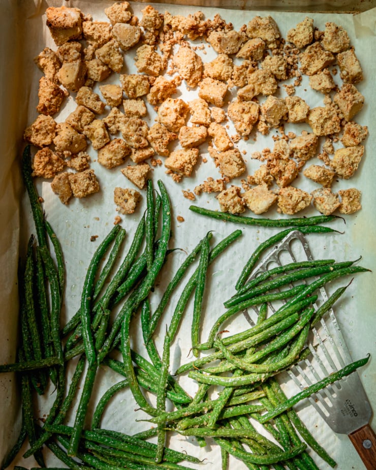 An overhead shot of roasted tofu and green beans on a parchment-lined sheet pan with a serving spatula nearby.