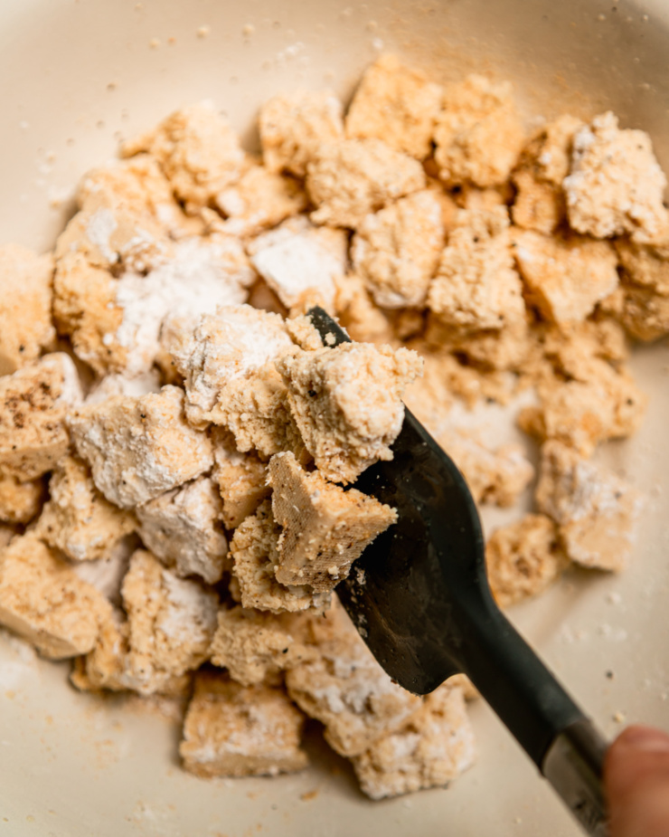 An overhead shot shows a spatula stirring up pieces of tofu with arrowroot starch, salt, pepper, and spices.