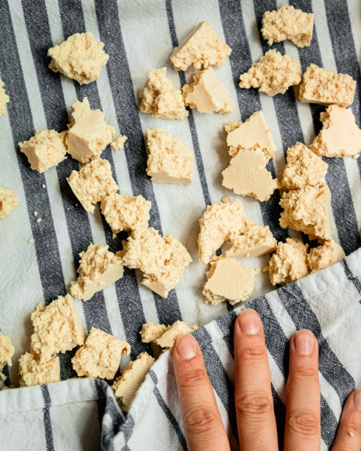 An up close, overhead shot of a hand pressing a towel into torn pieces of firm tofu.