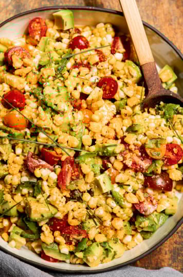 An up close, overhead shot shows a fresh corn, tomato & avocado salad in a shallow ceramic dish with a wooden spoon sticking out. The salad is topped with chopped chives and chopped corn nuts.