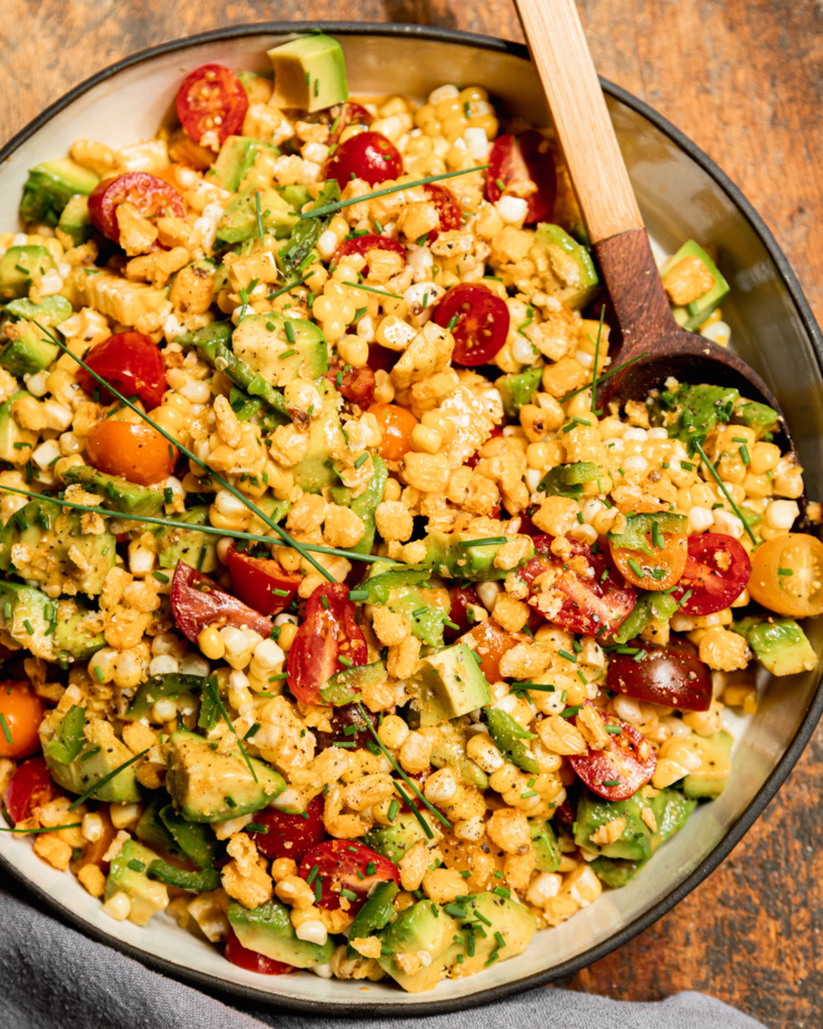 An up close, overhead shot shows a fresh corn, tomato & avocado salad in a shallow ceramic dish with a wooden spoon sticking out. The salad is topped with chopped chives and chopped corn nuts.