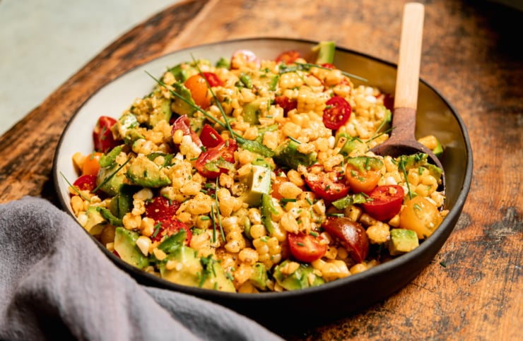 A 3/4 angle shot shows a fresh corn, tomato & avocado salad in a shallow ceramic dish with a wooden spoon sticking out. The salad is topped with chopped chives and chopped corn nuts. A blue linen napkin is nearby.