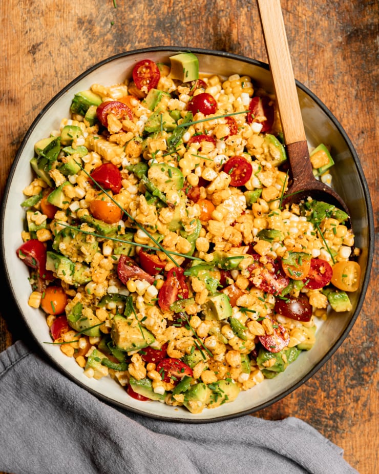 An overhead shot shows a fresh corn, tomato & avocado salad in a shallow ceramic dish with a wooden spoon sticking out. The salad is topped with chopped chives and chopped corn nuts.