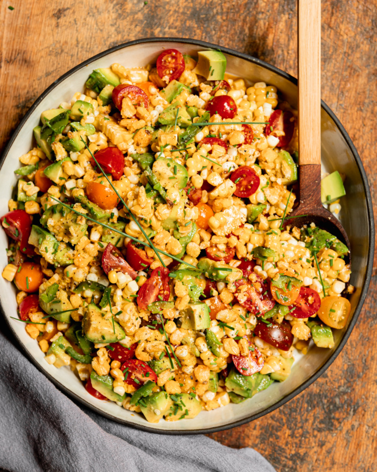 An overhead shot shows a fresh corn, tomato & avocado salad in a shallow ceramic dish with a wooden spoon sticking out. The salad is topped with chopped chives and chopped corn nuts.