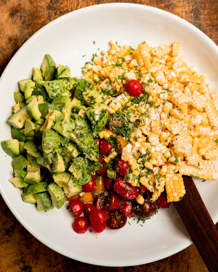 An overhead shot shows cooked corn, chives, halved small tomatoes, and diced avocado in a wide bowl.