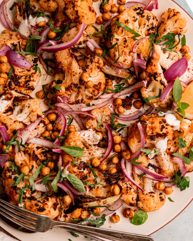 An up close, overhead shot of grilled cauliflower wedges on a platter over some yogurt tahini sauce. The cauliflower is topped with sliced sumac-pickled onions, chopped parsley, and crispy chickpeas.