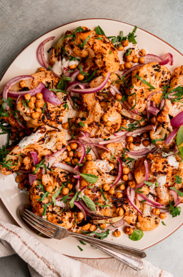 An overhead shot of grilled cauliflower wedges on a platter over some yogurt tahini sauce. The cauliflower is topped with sliced sumac-pickled onions, chopped parsley, and crispy chickpeas.