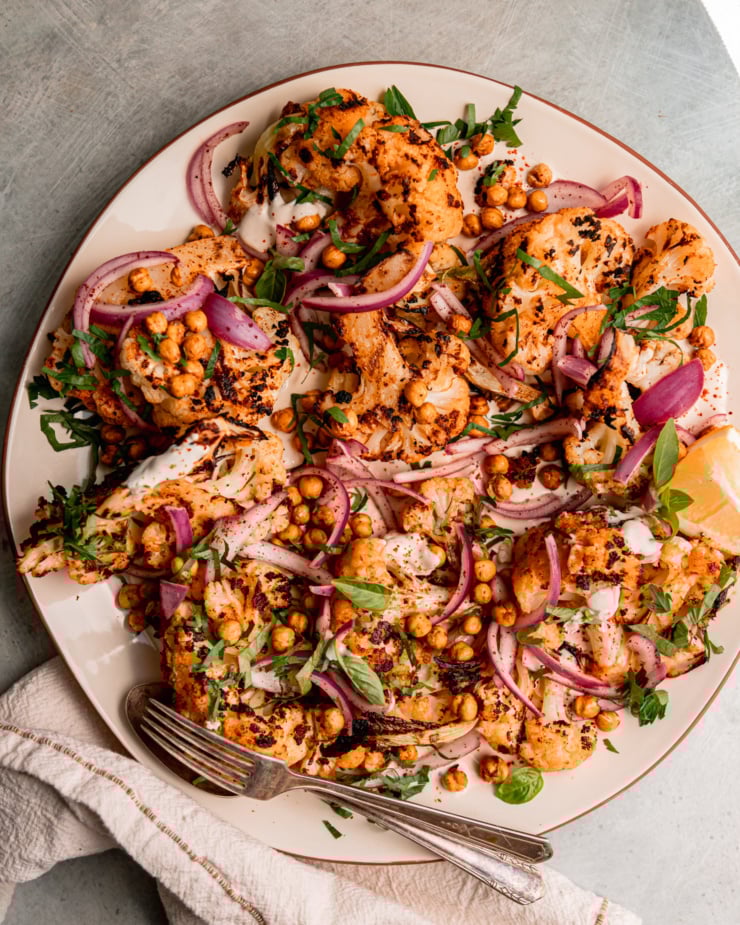 An overhead shot of grilled cauliflower wedges on a platter over some yogurt tahini sauce. The cauliflower is topped with sliced sumac-pickled onions, chopped parsley, and crispy chickpeas.