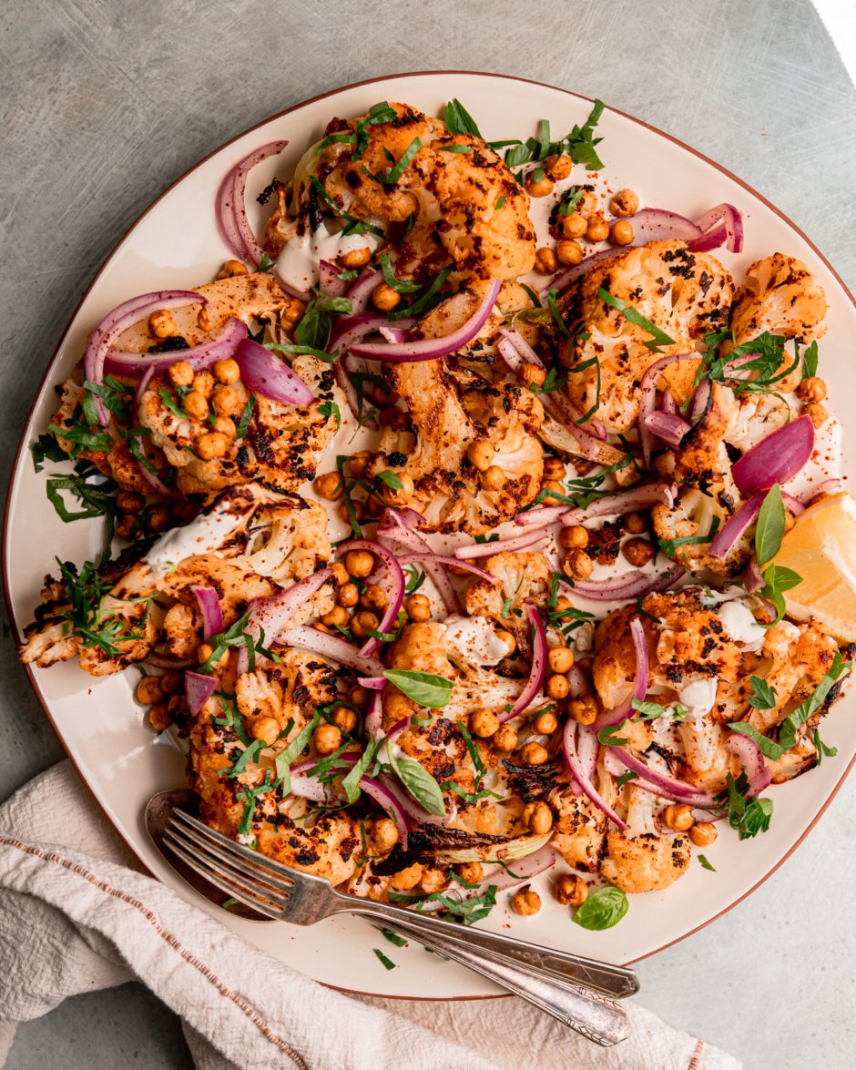 An overhead shot of grilled cauliflower wedges on a platter over some yogurt tahini sauce. The cauliflower is topped with sliced sumac-pickled onions, chopped parsley, and crispy chickpeas.