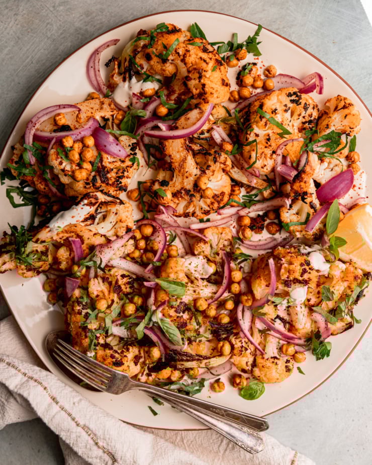 An overhead shot of grilled cauliflower wedges on a platter over some yogurt tahini sauce. The cauliflower is topped with sliced sumac-pickled onions, chopped parsley, and crispy chickpeas.