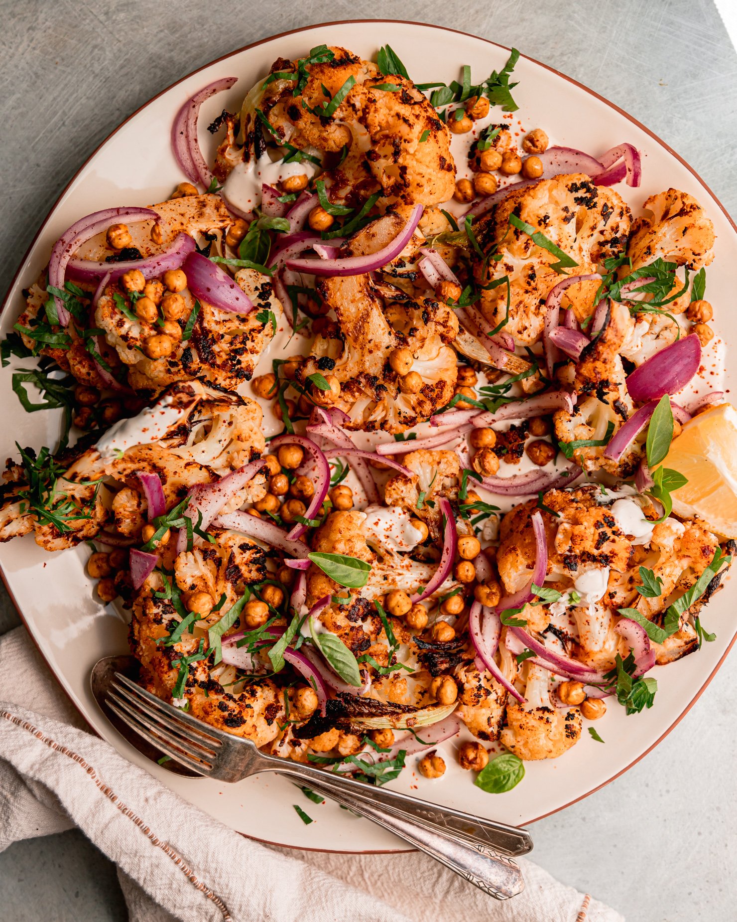 An overhead shot of grilled cauliflower wedges on a platter over some yogurt tahini sauce. The cauliflower is topped with sliced sumac-pickled onions, chopped parsley, and crispy chickpeas.