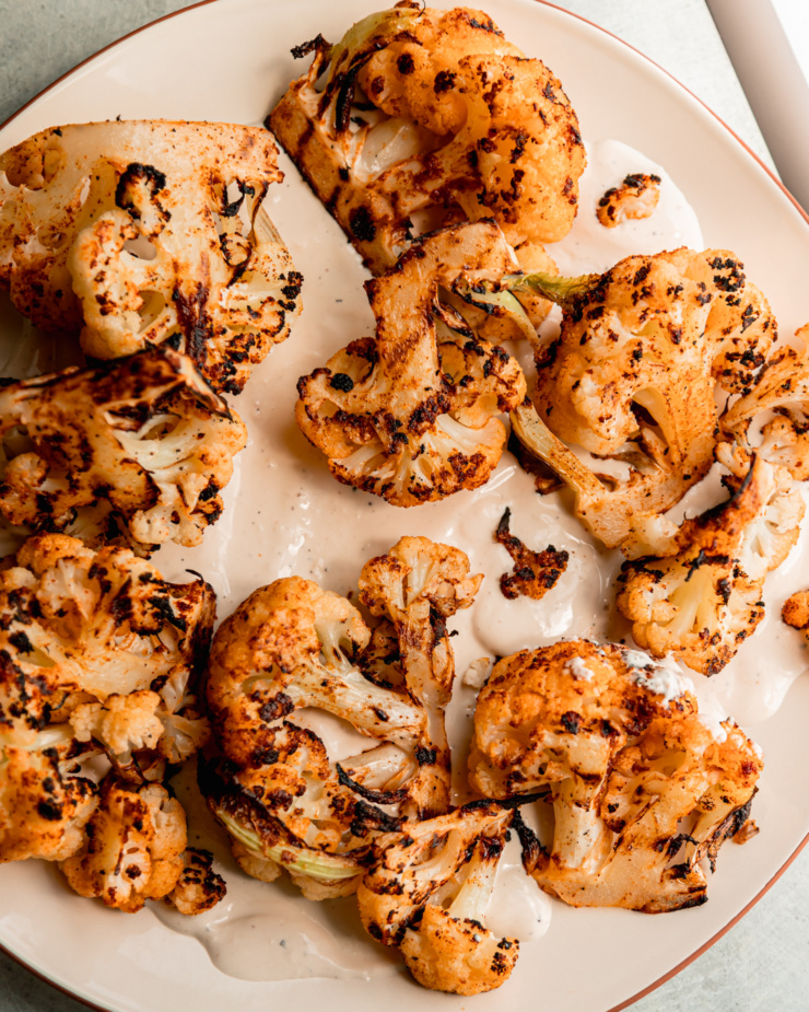 An overhead shot shows BBQ cauliflower pieces on top of a pool of creamy yogurt tahini sauce on a platter.