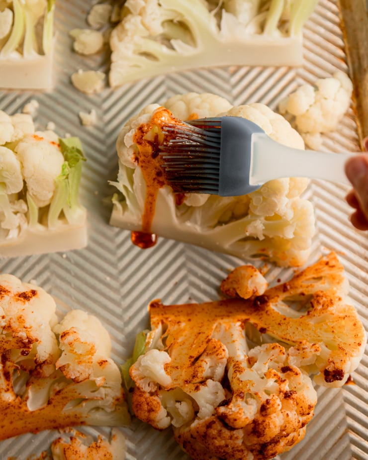 An overhead shot shows a brush applying a spice oil to steamed pieces of cauliflower on a baking sheet.