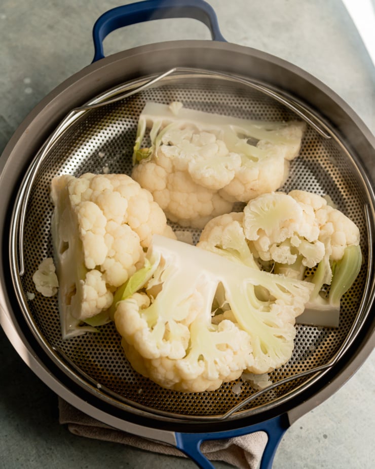 A slight 3/4 angle shot shows wedges of cauliflower being steamed in a basket inside of a deep skillet. There is steam coming off of the cauliflower.