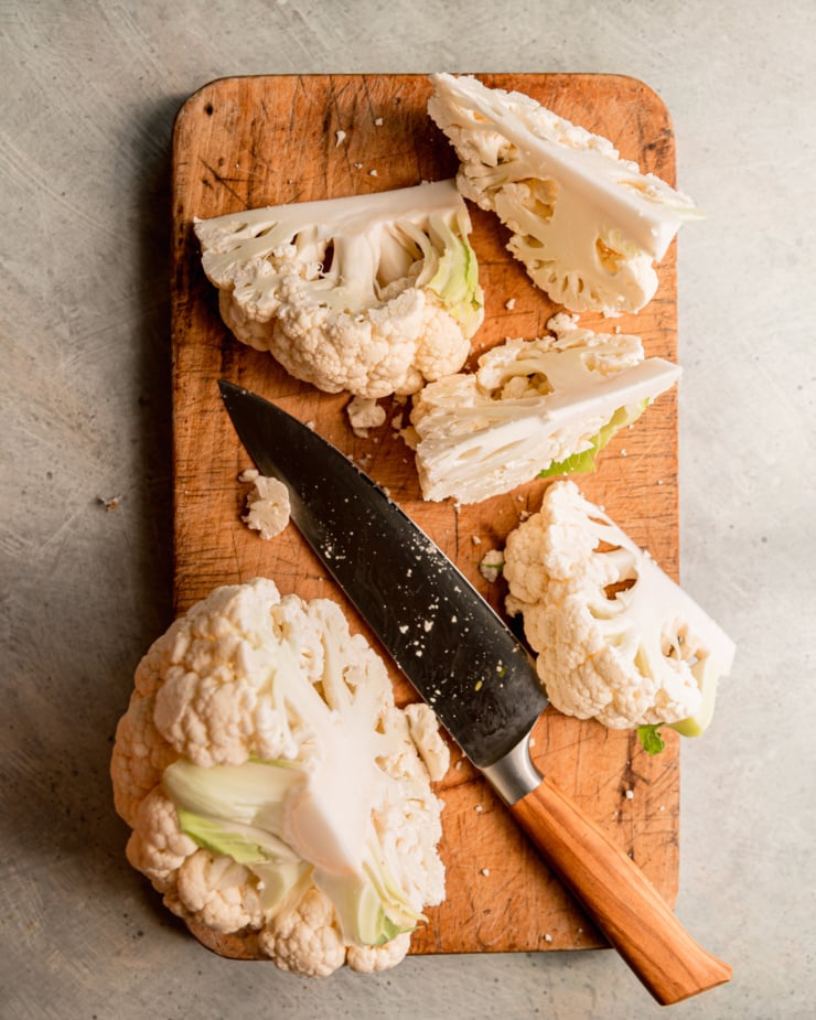 An overhead shot shows a head of cauliflower being cut into wedges on a worn wood cutting board.