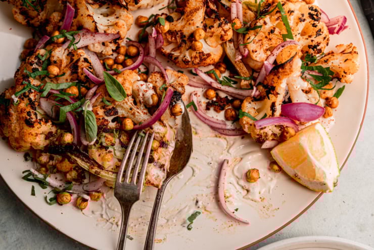 An up close, overhead shot of grilled cauliflower wedges on a platter over some yogurt tahini sauce. The cauliflower is topped with sliced sumac-pickled onions, chopped parsley, and crispy chickpeas. One serving of the cauliflower has been removed from the platter.