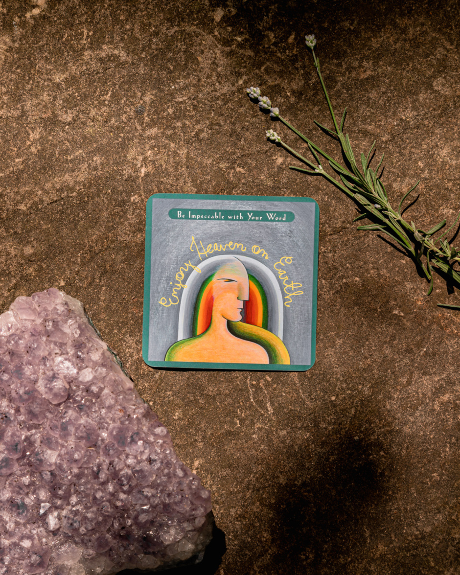 An overhead shot shows a card from The Four Agreements card deck that says "Enjoy Heaven on Earth." The card is next to an amethyst crystal and some fresh picked lavender buds.