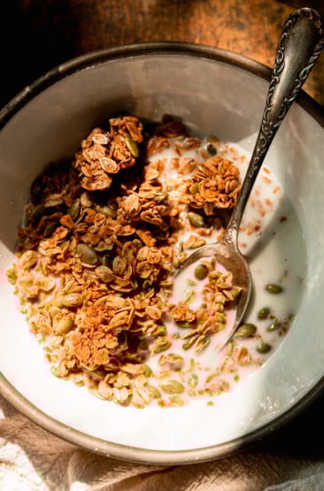 An overhead shot shows a ceramic bowl with pumpkin seed granola and some almond milk in bright sunlight.