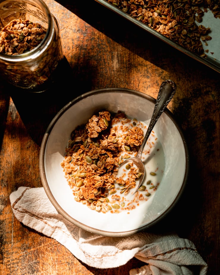 An overhead shot shows a ceramic bowl with pumpkin seed granola and some almond milk in bright sunlight. A jar and sheet pan of the granola is nearby.