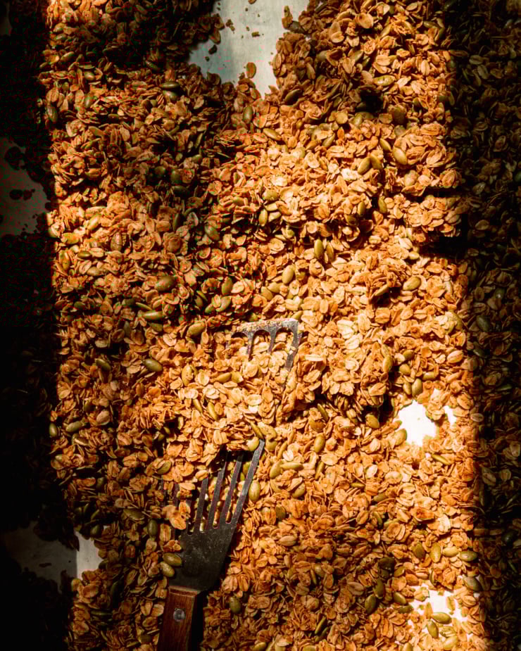 An overhead shot shows a baking sheet filled with golden pumpkin seed granola in direct evening light.