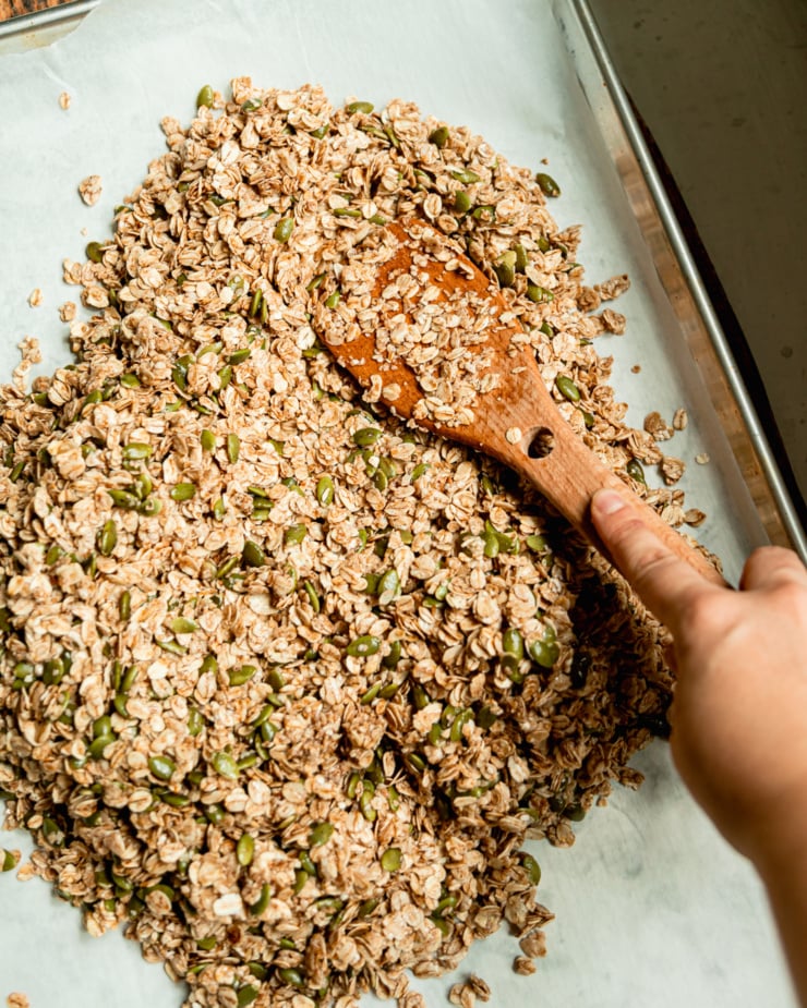 An overhead shot shows a hand using a wooden utensil to flatten out a raw granola mixture on a baking sheet lined with parchment paper.