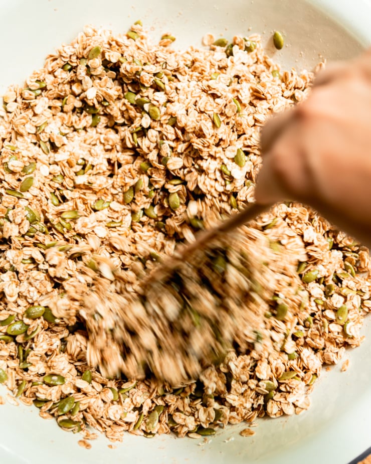 An overhead image shows a hand using a wooden utensil to stir up a pumpkin seed granola mixture.