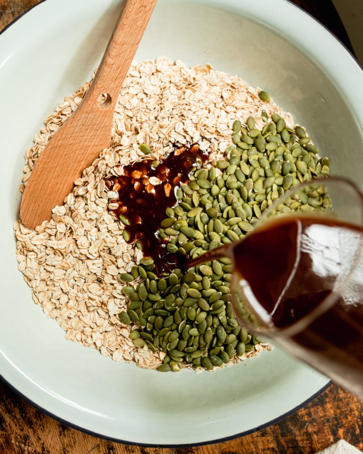 An overhead shot shows a maple syrup and coconut oil mixture being poured over toasted oats and pumpkin seeds in a large enamelware bowl.