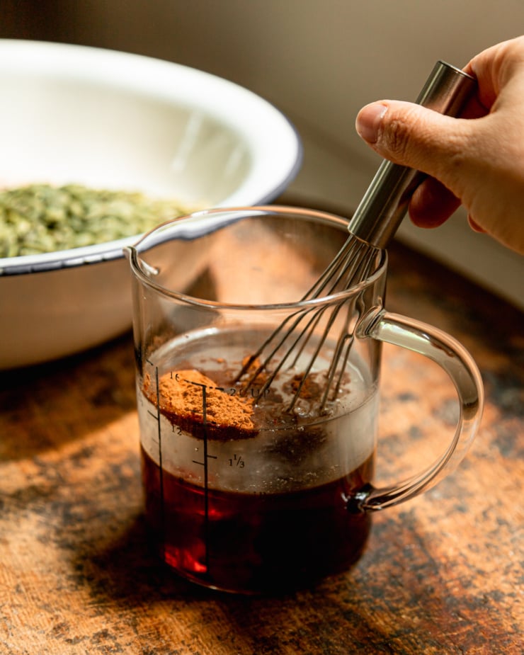 A head-on shot shows a hand using a whisk to combine coconut oil, maple syrup, cinnamon, salt, and vanilla extract in a glass measuring cup.