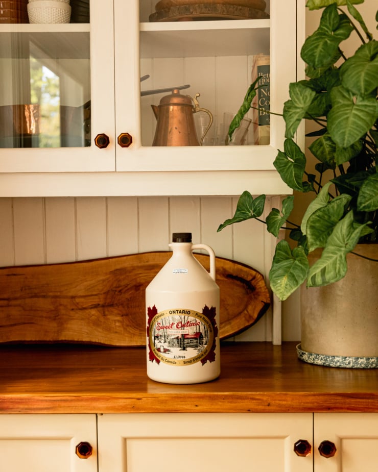 A head-on shot shows a large jug of maple syrup on a wooden counter underneath a cabinet. A large plant is nearby.