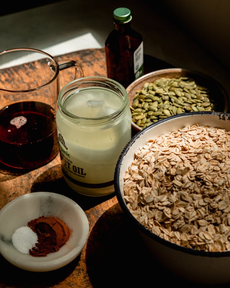 A 3/4 angle image shows the following ingredients on a worn wooden board: maple syrup, coconut oil, cinnamon, salt, rolled oats, pumpkin seeds, and vanilla extract.