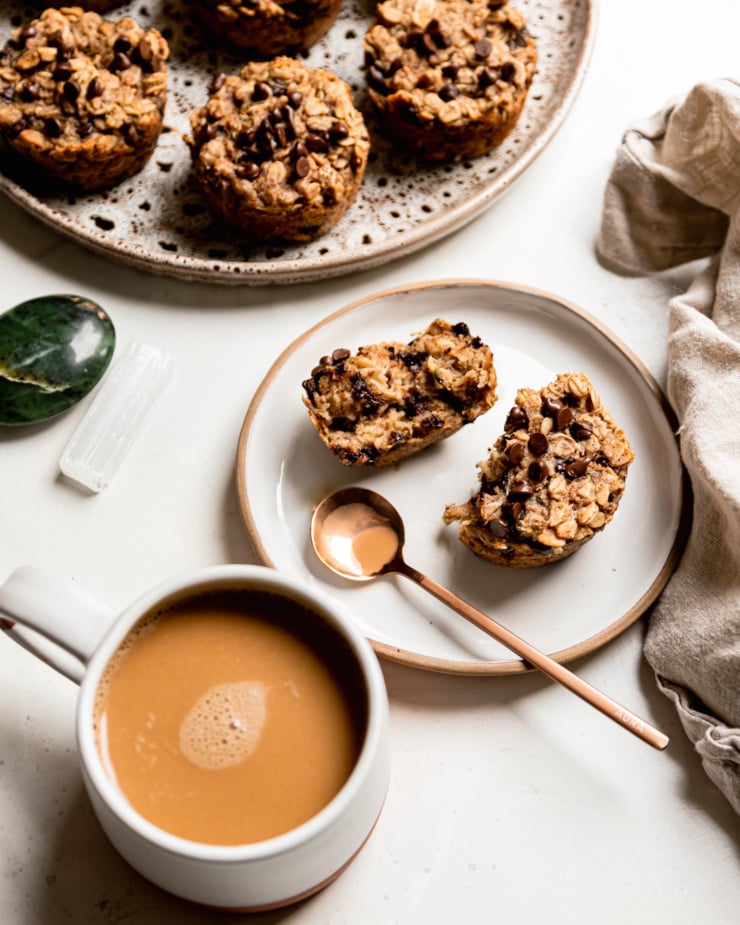 An overhead shot shows a split oatmeal cup on a plate next to a cup of coffee and a larger plate with more oat cups on it.