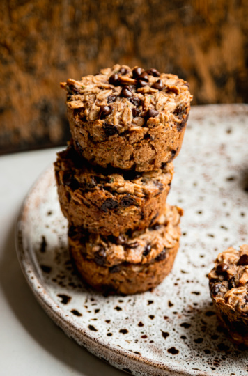 A slight 3/4 angle shot shows a stack of 3 zucchini baked oatmeal cups on a speckled plate. The cups have chocolate chips on top and a worn wooden board is in the background.