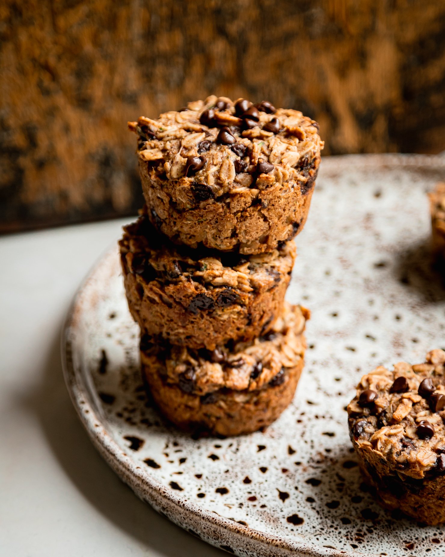 A slight 3/4 angle shot shows a stack of 3 zucchini baked oatmeal cups on a speckled plate. The cups have chocolate chips on top and a worn wooden board is in the background.