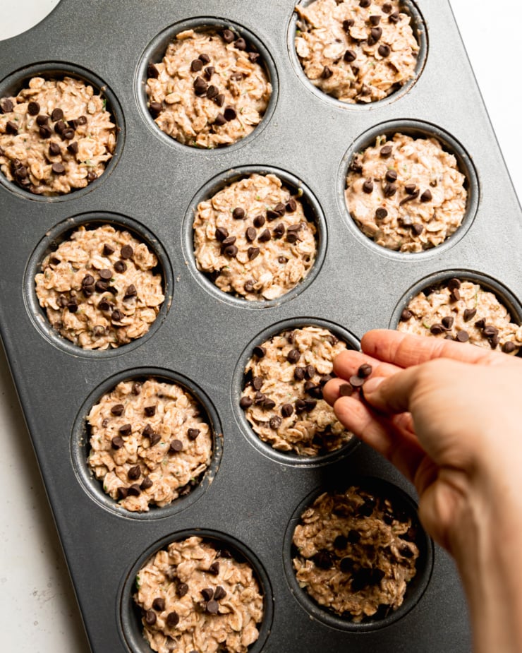An overhead shot shows a hand topping zucchini oatmeal cups with mini chocolate chips.