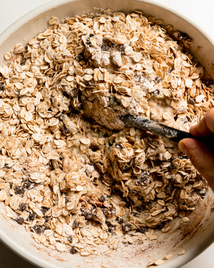 An overhead shot of a hand using a spatula to stir oats and oat flour into a batter.