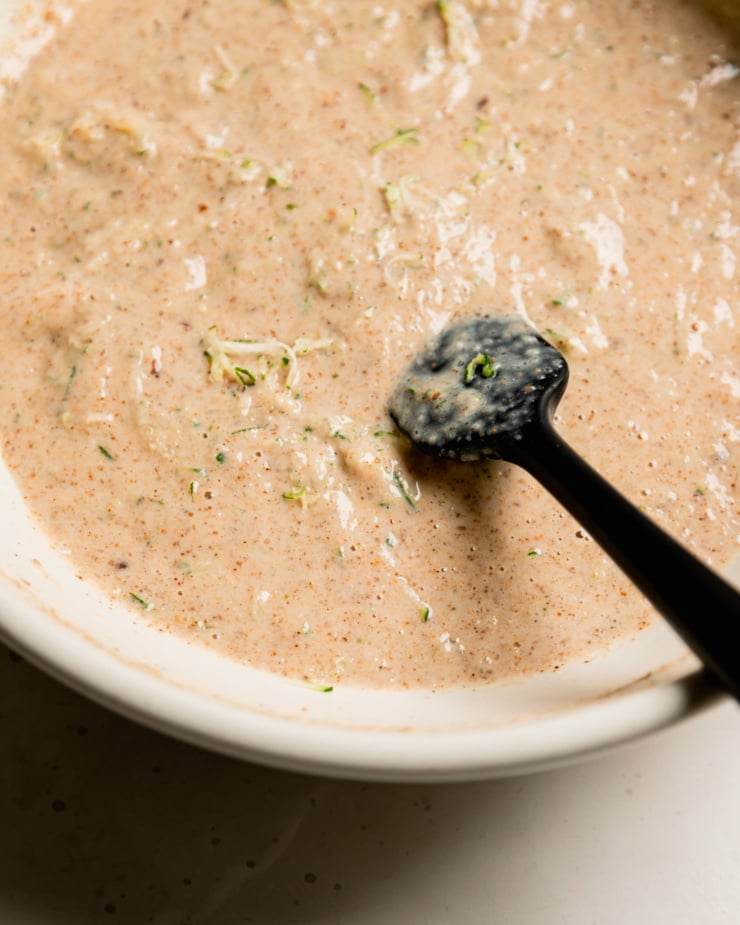 A 3/4 angle shot of a zucchini-flecked batter in a bowl with a spatula sticking out.