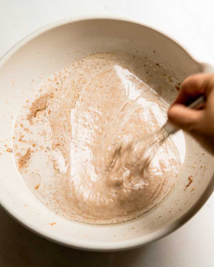 An overhead shot of a hand using a whisk to combine liquid ingredients in a large beige bowl.