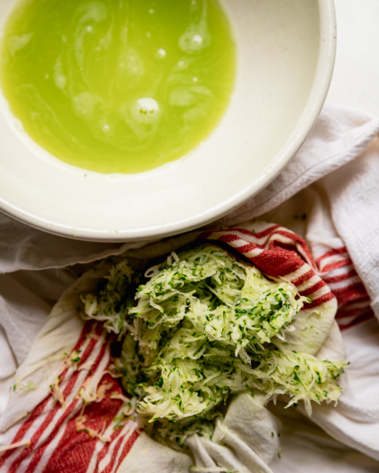 An overhead shot shows some wrung out grated zucchini and a bowl with the bright green zucchini liquid in it.